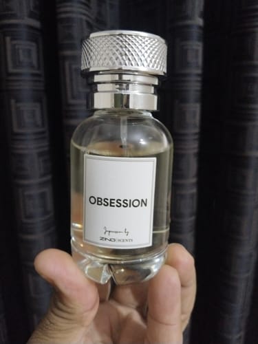 Customer's hand holding a clear bottle of Obsession perfume with a textured silver cap against a dark curtain.