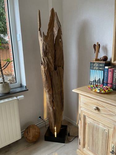 Ein Kunde zeigt die Teakholz Stehlampe ALMA, die aufgestellt in einer Zimmerecke sanft leuchtet.
