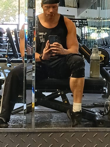 Customer taking a mirror selfie while seated on a gym bench, showing muscular arms and legs in a tank top.