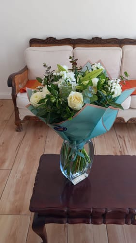 Customer's Classic White Bouquet with lush greenery, wrapped in teal paper and displayed in a clear glass vase on a wooden table.