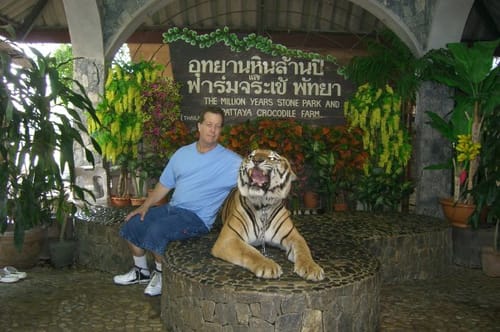 A reviewer for Tro+ Mune™ sits on a stone ledge next to a large tiger at an outdoor park.