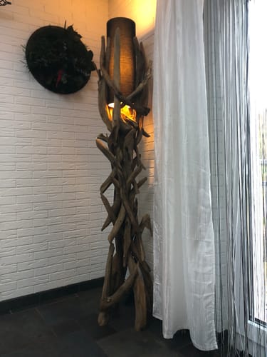 Ein Kunde zeigt die beleuchtete Treibholz-Stehlampe VILLA mit schwarzem Lampenschirm in einer Raumecke.