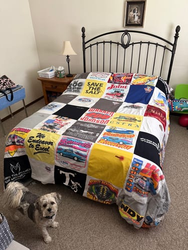Customer's finished T-Shirt Quilt with a motorsports theme spread on a bed, with a small dog standing in the foreground.