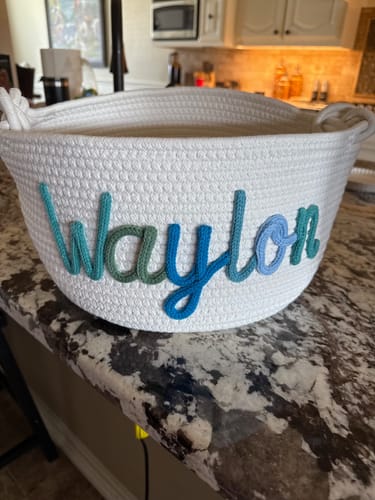 Customer's personalized white knitted basket with the name 'Waylon' in blue and green yarn on a kitchen counter.