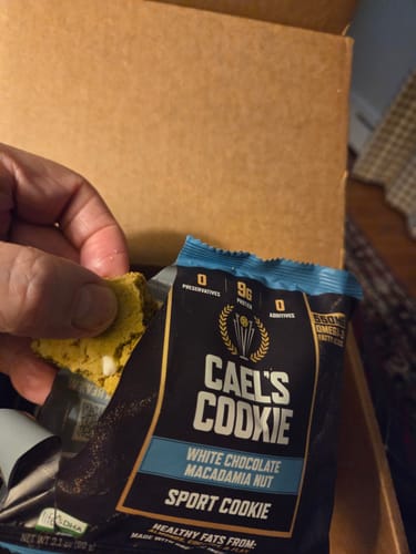 Customer's hand holding a broken Cael’s White Chocolate Macadamia Nut cookie over the open packaging.