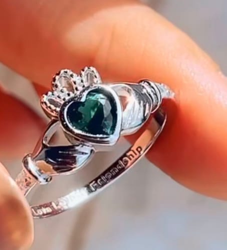 Close-up of a reviewer's hand holding a silver Claddagh ring with a green, heart-shaped stone.