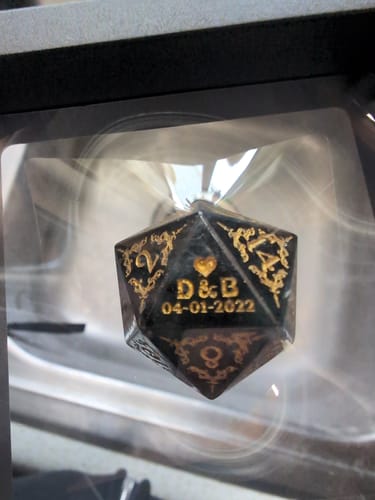 Close-up of a customer's black D20 die with ornate gold engraving, showing the personalized face with 'D & B' and '04-01-2022'.