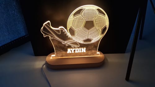 Recensent toont de verlichte, gepersonaliseerde 3d voetballamp met de naam 'AYDIN' op een houten voet.