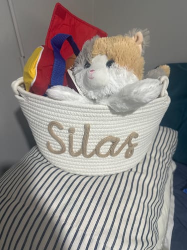 Customer's white personalized knitted basket with the name 'Silas' in beige yarn, filled with a stuffed cat and other toys.