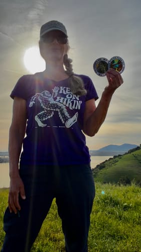 Reviewer wearing the purple Keep On Hikin' tee while standing on a sunny, grassy hill near water.