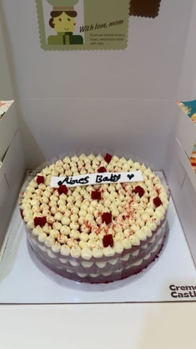 Customer's Red Velvet cake in its box, topped with piped white frosting, red sprinkles, and a personalized message.