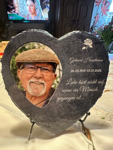 Ein Kunde zeigt das personalisierte Schieferherz mit einem Foto und graviertem Text, das auf einem Ständer auf einem Tisch steht.