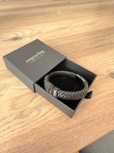 Customer's black braided bracelet with steel details, shown inside its open grey gift box on a wooden table.
