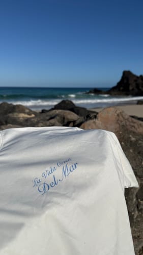 Customer's white Del Mar T-Shirt with blue text, displayed on rocks at a sunny beach.