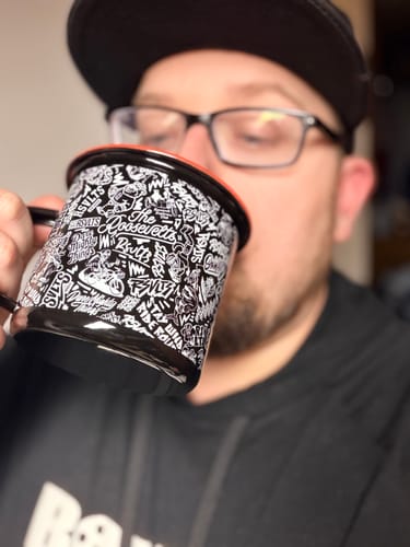A close-up of a reviewer sipping from the black enamel "Outpost" mug, showing its detailed white pattern.