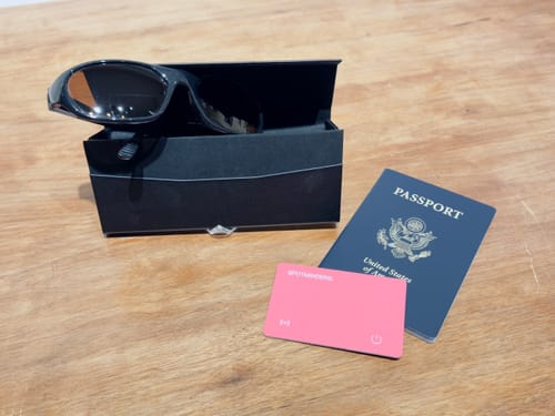 Customer's black Spotminders glasses case holding sunglasses, next to a passport and a pink tracking card.