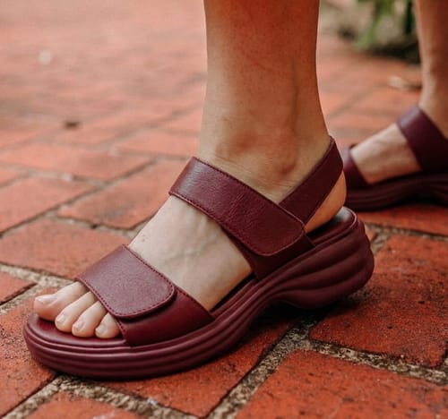 A reviewer's foot wearing the burgundy Modesty sandal, showing its thick platform sole and adjustable straps on a brick path.