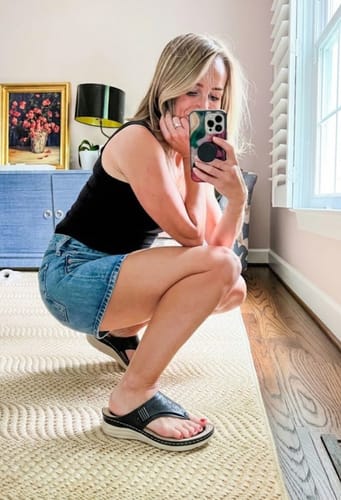 Reviewer crouching in a room while wearing the black Grace wedge sandals with a denim skirt.