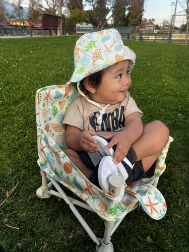 Customer's toddler sits in the Sailor portable booster chair on a grassy lawn wearing a matching hat.