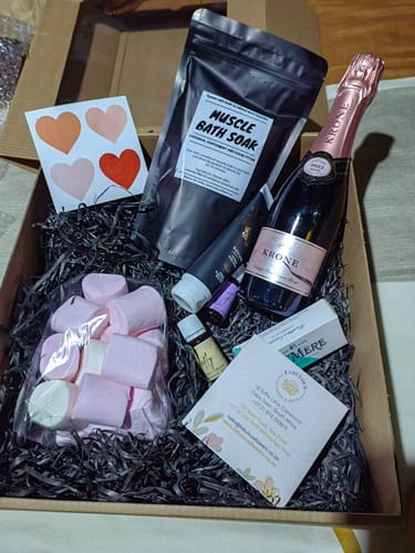 Customer's open Pamper and Indulge Gift Box showing rosé, bath soak, marshmallows, and lotion nestled in black paper.