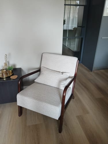 Recensent toont de beige Wise fauteuil met een donker walnoot frame in een woonkamer.