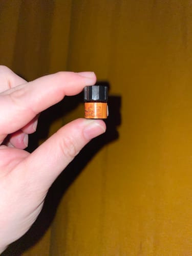 Reviewer's hand holding a tiny sample vial of the orange Ember perfume against a warm background.