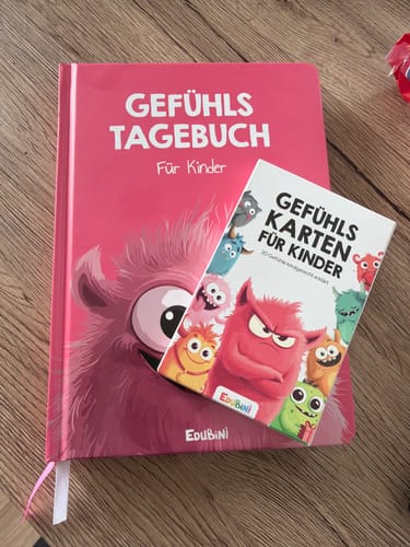 Ein Rezensent zeigt das rosafarbene Gefühlstagebuch für Kinder mit einem Satz Gefühlskarten auf einem Holztisch.