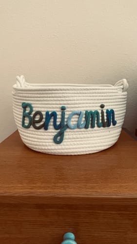 Customer's white knitted basket sitting on a dresser, personalized with the name 'Benjamin' in multicolor yarn.