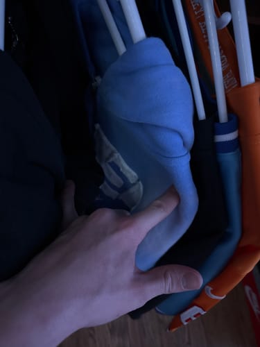 Close-up of a customer's hand pointing to the fabric of a light blue SEEK ZIPUP hoodie hanging in a closet.
