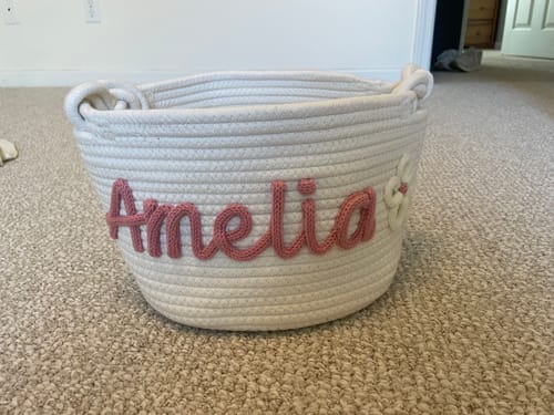 Customer's white woven basket personalized with the name 'Amelia' in pink yarn and a small flower.
