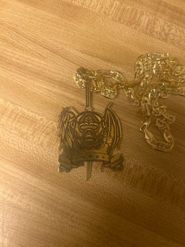 Customer's custom gold pendant necklace with a detailed winged skull and sword design, shown on a wooden surface.