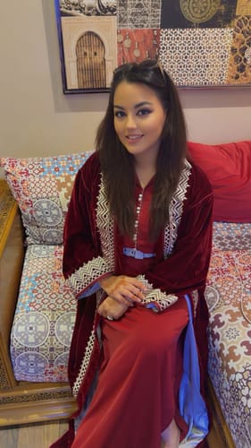 Customer wearing the red velvet Kovas dress, showing its detailed silver embroidery while sitting indoors.
