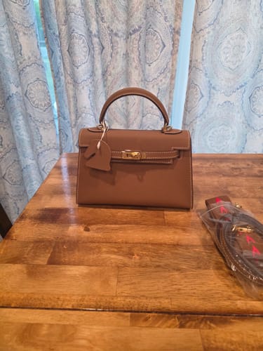 Reviewer's brown structured Kally Leather Bag sitting on a wooden table next to its packaged shoulder strap.