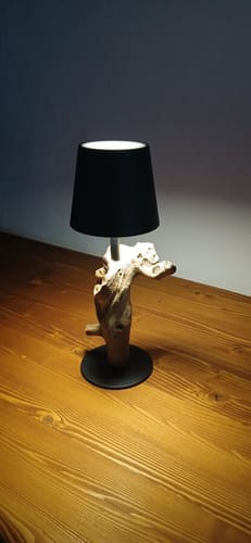 Ein Kunde zeigt die eingeschaltete SERA Tischlampe mit schwarzem Schirm und Treibholzfuß auf einem Holztisch.
