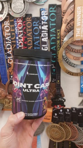 Reviewer holding a black tub of Joint Care Ultra in front of a collection of race medals.