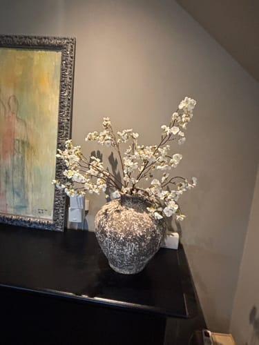 Recensent toont de witte Zijden Bloesem in een rustieke, getextureerde vaas op een donker meubel.
