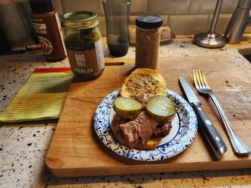 Customer's prime rib sandwich on a cutting board, topped with two large Big Dill Pickles, with the jar in the background.