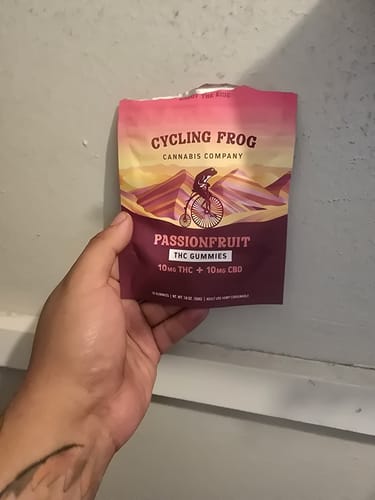 Customer holding a colorful pouch of Cycling Frog Passionfruit Gummies against a plain wall.