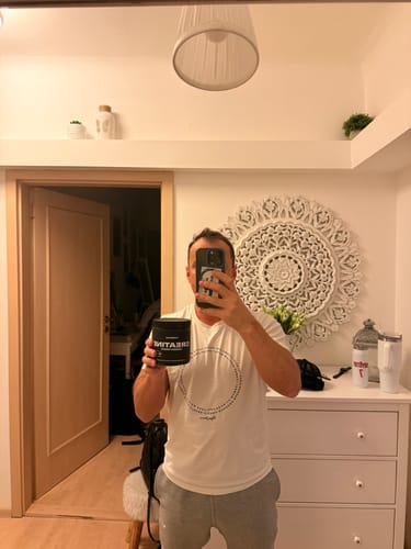 Customer taking a mirror selfie in their room, holding up a black tub of Creatine Monohydrate.