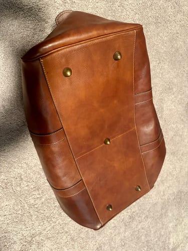 Reviewer shows the bottom of the brown leather Wanderer Duffel, highlighting the five protective metal feet.