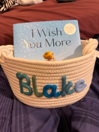 Customer's personalized knitted basket with the name 'Blake' in multi-colored yarn, shown holding a children's book.