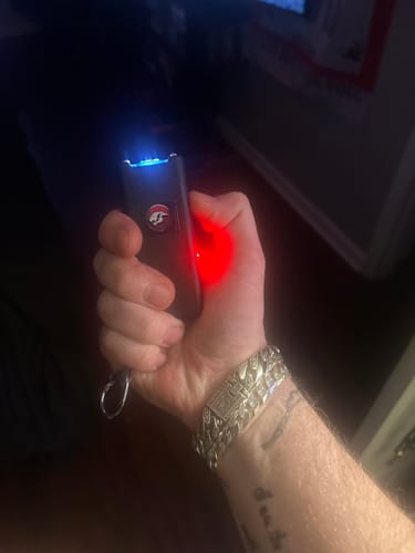Reviewer holding the activated Black Hornet Stun Gun, showing its blue electrical arc and red indicator light in a dark room.