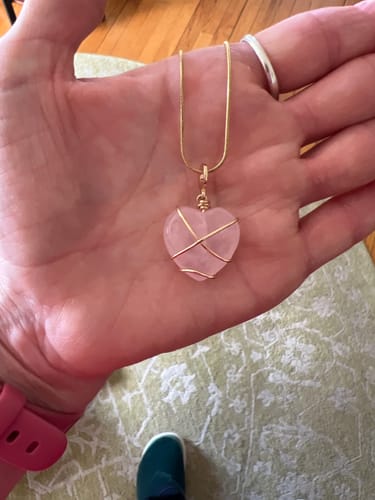 Reviewer's hand holding a heart-shaped pink crystal pendant with gold wire wrapping on a gold chain.