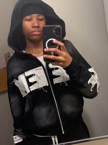 Reviewer in a mirror selfie wearing the black SEEK ZIPUP, showing its fit and distressed white embroidery.