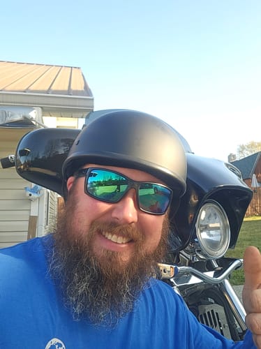 Reviewer with a beard smiling and giving a thumbs-up in front of a motorcycle; the neck gaiter is not visible.