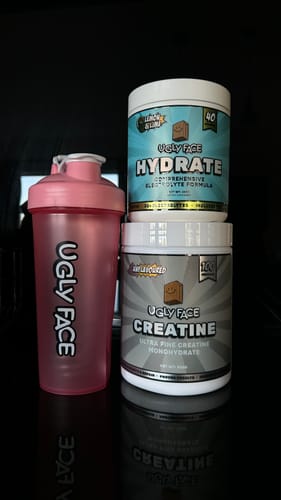 Reviewer's tub of Ugly Face Creatine Monohydrate shown on a reflective surface next to a pink shaker cup.