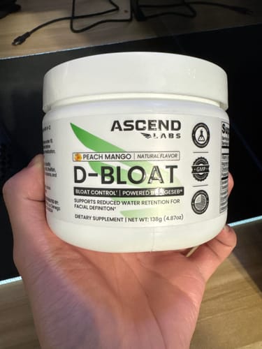 Reviewer's hand holding a white tub of D-BLOAT, a peach mango flavored supplement powder.