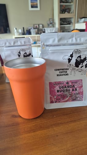 Customer's pouch of Uganda Bugisu AA coffee next to an orange tumbler on a wooden table.