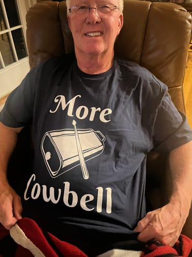 A smiling reviewer sits in a chair wearing the dark 'More Cowbell' T-shirt, showing its loose fit.
