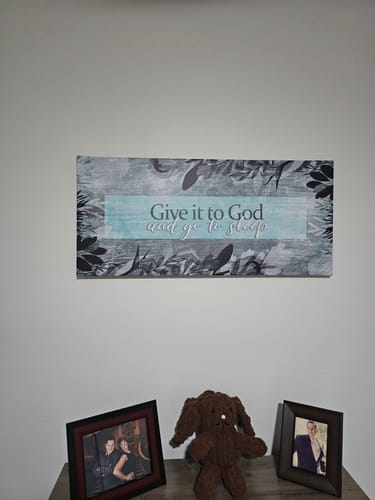 Customer's 'Give it to God' wall art hanging above a table with framed pictures in a bedroom.